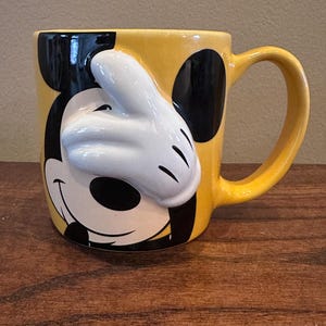 May include: Yellow ceramic mug featuring Mickey Mouse's face and a white 3D glove. The mug has a black ear on each side and a yellow handle. A fun, collectible Disney-themed mug.