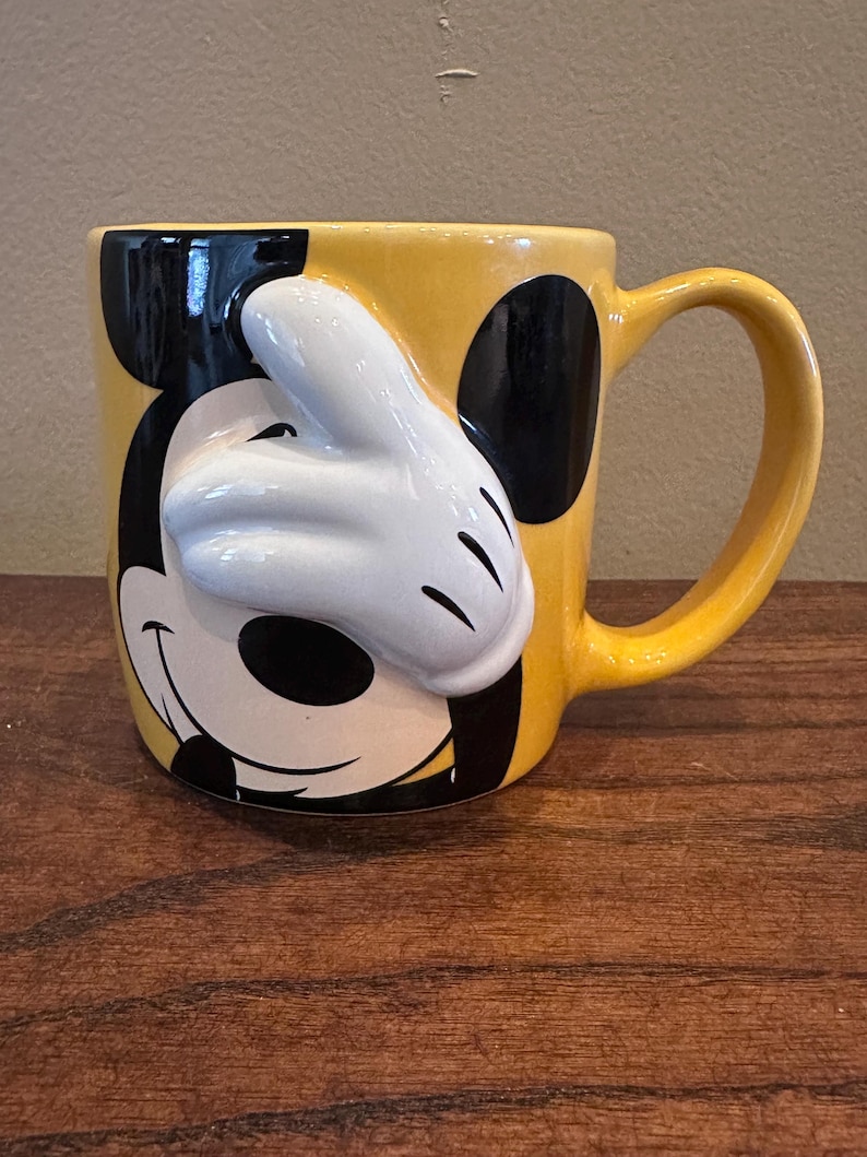 May include: Yellow ceramic mug featuring Mickey Mouse's face and a white 3D glove. The mug has a black ear on each side and a yellow handle. A fun, collectible Disney-themed mug.