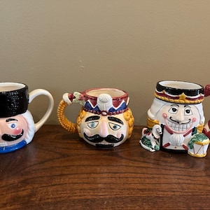 Op de afbeelding: Drie keramische notenkraker mokken met gedetailleerde gezichten en handvatten. De mokken hebben een zwarte hoed, een kroon en een witte pruik. De mokken zijn beschilderd met rode, blauwe en gouden accenten. De mokken staan op een houten oppervlak.