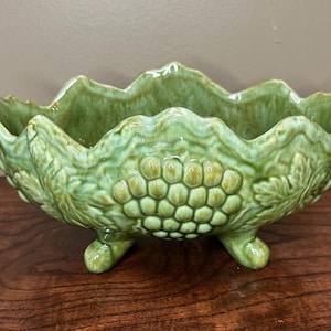 May include: A green ceramic bowl with a scalloped rim and three small feet. The bowl is decorated with grape clusters and leaves, and has a glossy finish. The bowl is sitting on a wooden surface.