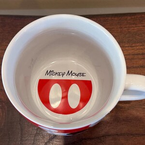 May include: White ceramic mug with a red and white Mickey Mouse design inside. The mug features the words "Mickey Mouse" in black script above the design. The mug has a white handle and a red band around the bottom.