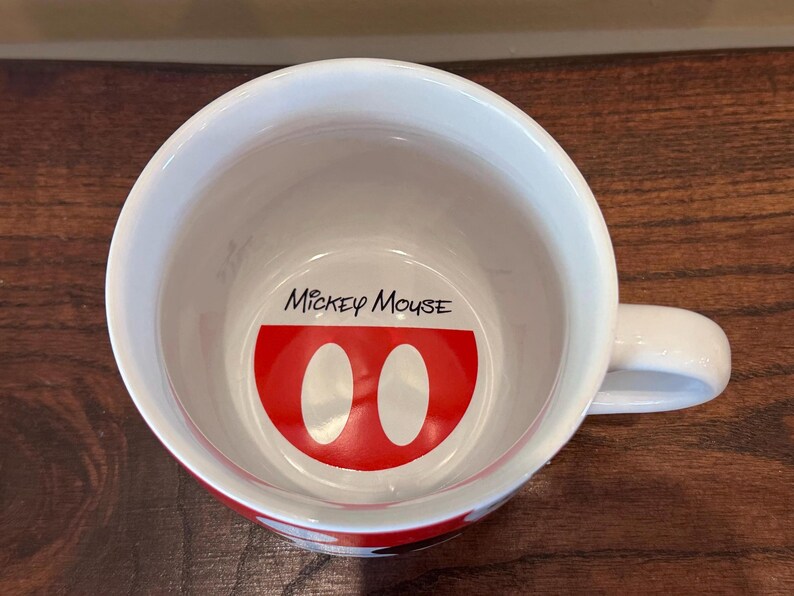 May include: White ceramic mug with a red and white Mickey Mouse design inside. The mug features the words "Mickey Mouse" in black script above the design. The mug has a white handle and a red band around the bottom.