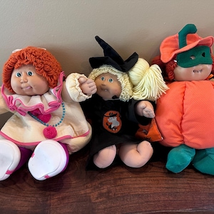 May include: Three Cabbage Patch Kids dolls dressed in costumes. The left doll has red yarn hair and a clown outfit. The centre doll has blonde yarn hair and a black witch costume. The right doll wears a pumpkin suit with a green hat and mask.