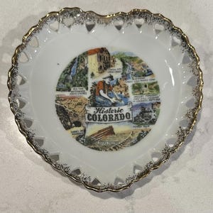 Könnte beinhalten: Herzförmige Zierplatte mit weißem Grund und goldfarbenem, gewelltem Rand. Die Mitte zeigt eine farbenfrohe Illustration von Wahrzeichen Colorados, einschließlich des Textes "Historic Colorado".