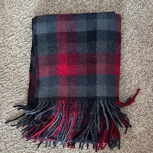 Puede incluir: Una manta plegada con un diseño de cuadros rojos, negros y grises. La manta presenta un patrón de cuadros clásico y un borde con flecos. El material parece suave y tejido, ideal para abrigarse.