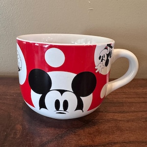 May include: A large ceramic mug featuring Mickey Mouse. The mug has a red background with white polka dots and a white handle. Mickey's face is prominently displayed on the front, with additional character illustrations around the mug.