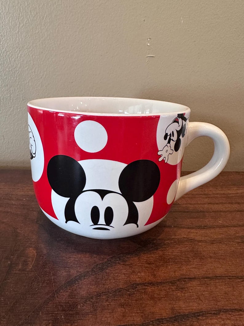 May include: A large ceramic mug featuring Mickey Mouse. The mug has a red background with white polka dots and a white handle. Mickey's face is prominently displayed on the front, with additional character illustrations around the mug.