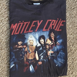 以下が含まれることがあります： Mötley Crüeのバンドロゴがプリントされた黒の半袖Tシャツ。赤い文字でバンド名が記され、その下にはスタッズやレザーをあしらったダークなステージ衣装を身にまとった4人のバンドメンバーの写真がデザインされています。