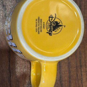 May include: Yellow ceramic Disney Store mug with a rounded handle. The bottom of the mug features the Disney Store logo and text that reads "MADE IN THAILAND" and "FABRIQUE EN THAILANDE".