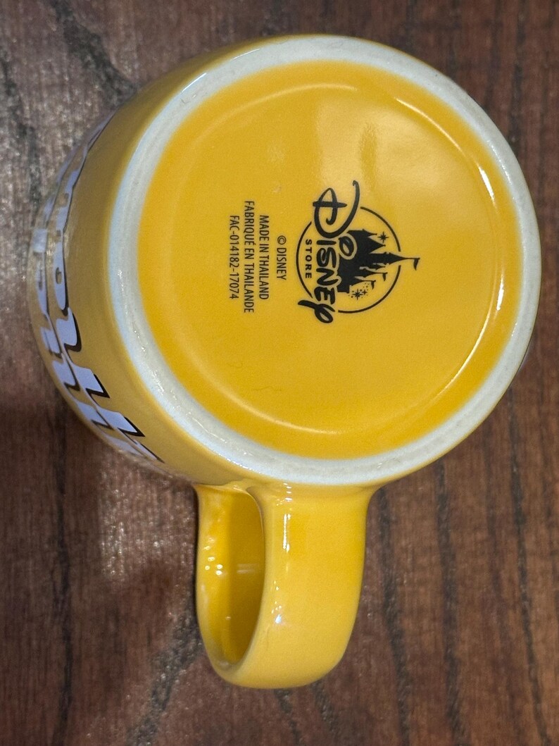 May include: Yellow ceramic Disney Store mug with a rounded handle. The bottom of the mug features the Disney Store logo and text that reads "MADE IN THAILAND" and "FABRIQUE EN THAILANDE".
