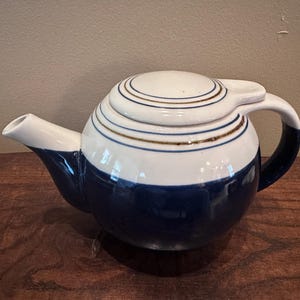 May include: A round ceramic teapot featuring a two-tone design with a deep navy blue base and a white upper section. The lid and the top of the body are decorated with thin, concentric brown circular lines. It includes a curved spout and handle.