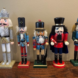 Könnte beinhalten: Fünf dekorative Nussknacker aus Holz in verschiedenen Designs. Die Nussknacker sind in Silber, Blau, Rot und Grün bemalt und weisen detaillierte Gesichtszüge und verzierte Uniformen auf. Jede Figur steht auf einem rechteckigen Sockel, ideal für die Weihnachtsdekoration.
