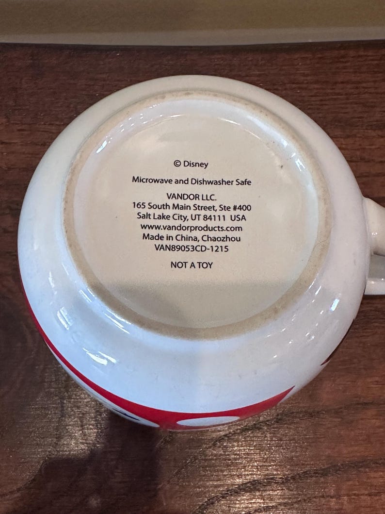 May include: White ceramic mug with a red and black design. The bottom of the mug features text including "&copy; Disney", "Microwave and Dishwasher Safe", and "NOT A TOY".