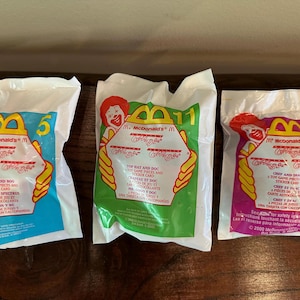 May include: Three sealed McDonald's Happy Meal toys in white plastic bags. The bags have colorful graphics with the McDonald's logo, Ronald McDonald, and the number 5, 11, and 7. The bags are on a wooden surface.