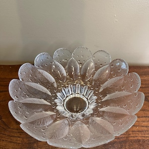May include: Clear glass serving dish in the shape of a flower. The dish has a central circular area and petal-shaped sections around the edge. The petals have a textured surface with small bubble-like details and leaf-like patterns.