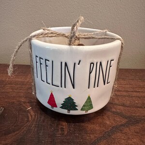 May include: A white ceramic mug with the black text FEELIN' PINE printed on the front. Below the text are three small illustrated Christmas trees in red, dark green, and light green. A piece of brown twine is wrapped around the top rim.
