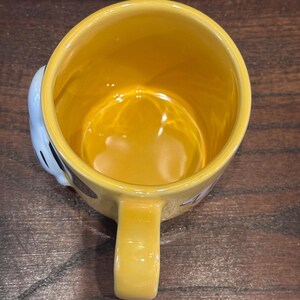 May include: A yellow ceramic mug with a matching handle. The interior of the mug is yellow, and the exterior features a cartoon design. The mug is set against a wooden surface.
