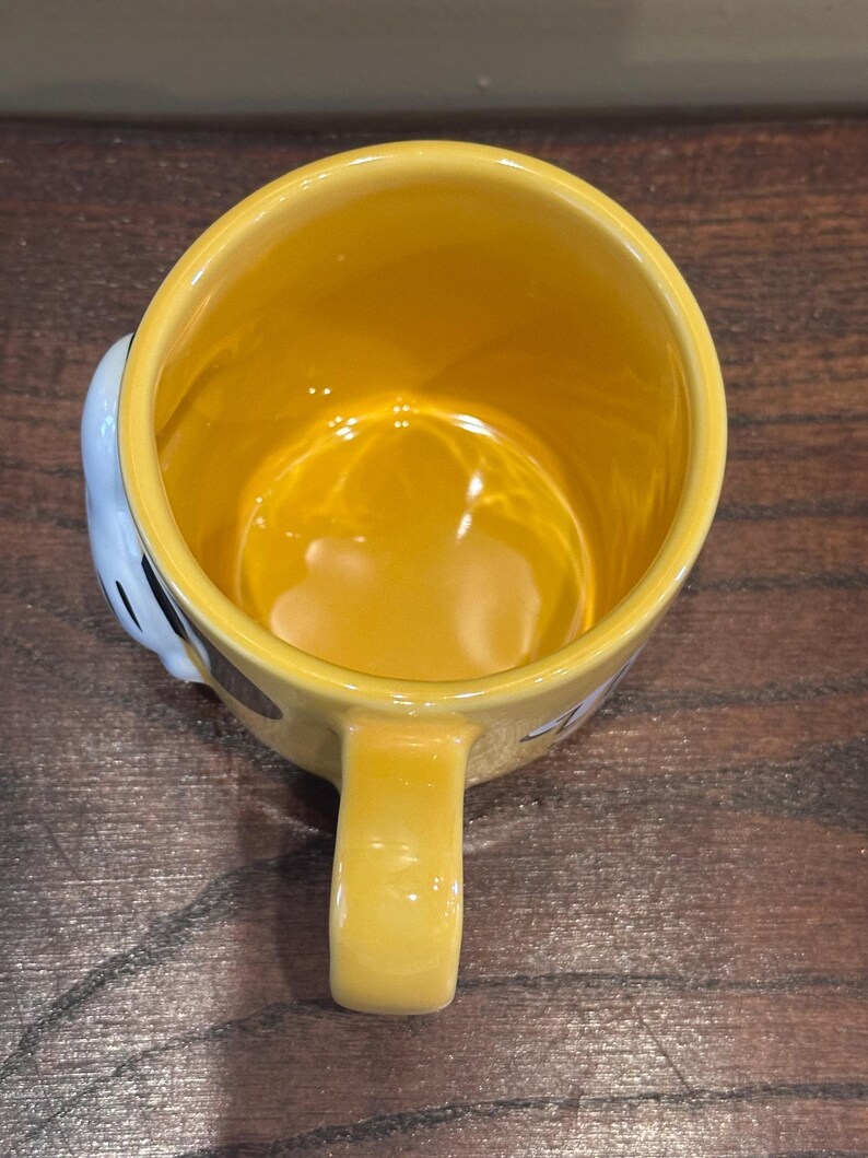 May include: A yellow ceramic mug with a matching handle. The interior of the mug is yellow, and the exterior features a cartoon design. The mug is set against a wooden surface.