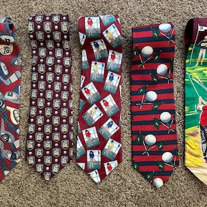 May include: Five golf-themed neckties in various designs. One tie features golf clubs and balls, another has a repeating pattern of golf shields, and a third displays golfers. The remaining ties have golf balls and flags.