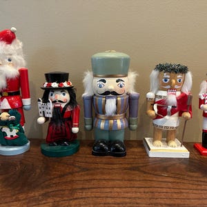 Könnte beinhalten: Eine Sammlung von sechs dekorativen Nussknackern aus Holz in verschiedenen Designs. Die Nussknacker sind in leuchtenden Farben bemalt, darunter Rot, Grün und Blau. Einige halten Requisiten wie einen Baum, einen Sack oder eine Zuckerstange. Sie stehen auf einer Holzoberfläche.