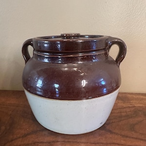 May include: A brown and white glazed ceramic crock with two handles and a lid. The crock is a traditional style with a rounded body and a wide mouth.