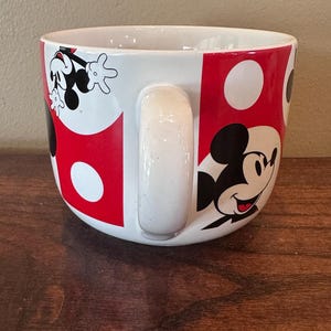 May include: White ceramic mug with a large handle, featuring a design of Mickey Mouse. The mug has a red and white polka dot pattern with Mickey Mouse illustrations. The mug is sitting on a wooden surface.