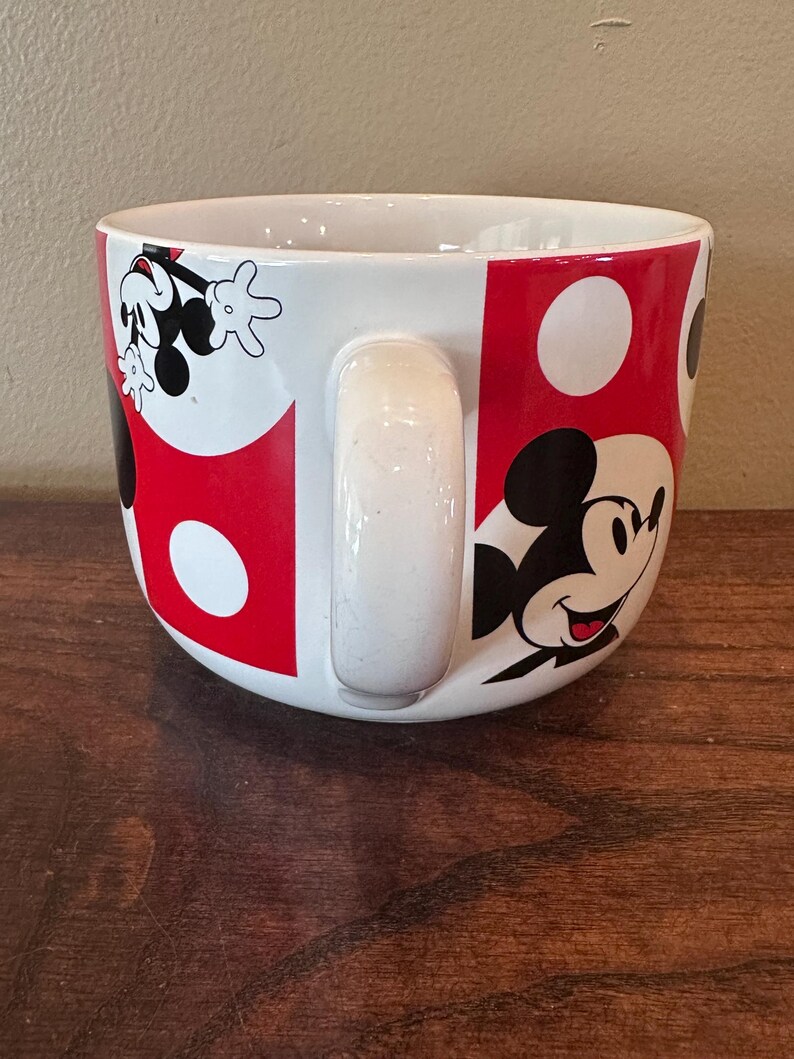May include: White ceramic mug with a large handle, featuring a design of Mickey Mouse. The mug has a red and white polka dot pattern with Mickey Mouse illustrations. The mug is sitting on a wooden surface.