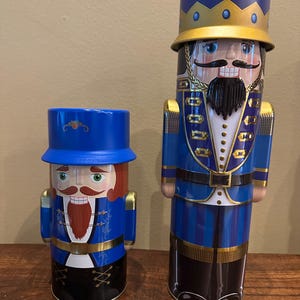 May include: Two decorative nutcracker figures. The figures are cylindrical with blue and gold accents. The larger nutcracker has a blue crown and a black beard. The smaller nutcracker has a red beard and a blue hat.