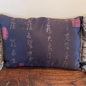 Peut inclure: Un coussin rectangulaire bleu marine avec des inscriptions et des tampons rouges. Le coussin a une garniture à franges aux extrémités. L'écriture est de couleur gris clair. Le coussin est sur une surface en bois.