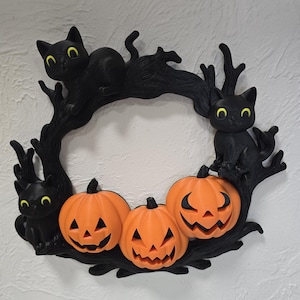 May include: A Halloween wreath featuring three orange jack-o'-lanterns with carved faces and three black cat figures with yellow eyes. The pumpkins and cats are set against a black, tree-like frame.