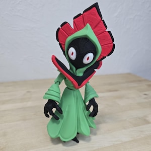 May include: A green action figure with a red and black floral headpiece and a black face. The figure has a green robe and black hands. The figure is posed on a wooden surface.