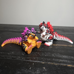 May include: Two colorful, stylized figurines of a Godzilla-like creature. One is orange and purple, the other is red, black, and white. Both have spiky dorsal fins and detailed features, sitting on a dark wooden surface.