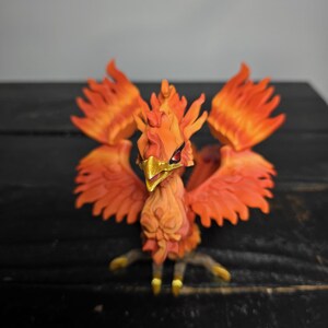 May include: A detailed figurine of a phoenix, featuring vibrant orange and red feathers. The bird has a golden beak and talons, with a detailed, textured body. The wings are spread, and the figure is set against a dark background.
