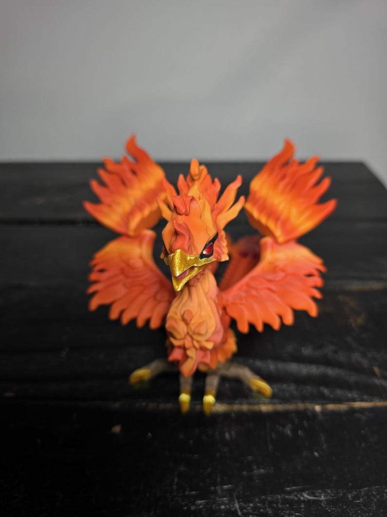 May include: A detailed figurine of a phoenix, featuring vibrant orange and red feathers. The bird has a golden beak and talons, with a detailed, textured body. The wings are spread, and the figure is set against a dark background.