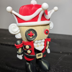 May include: A whimsical figurine of a nutcracker soldier. The figure has a red and white crown, a red jacket with black accents, and black boots. The face is a light tan color with a button eye and a toothy grin. The figurine is standing upright.
