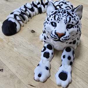 May include: A white and black 3D printed snow leopard figurine with a segmented tail. The figurine has a realistic appearance with black spots and a detailed face.