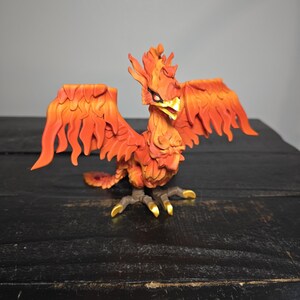 May include: A detailed figurine of a phoenix with outstretched wings. The bird is predominantly orange and red, with yellow accents on its beak and talons. The phoenix is posed on a dark surface.