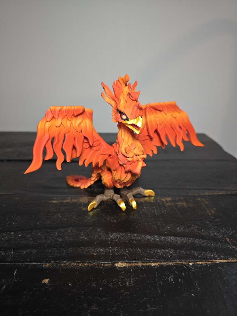 May include: A detailed figurine of a phoenix with outstretched wings. The bird is predominantly orange and red, with yellow accents on its beak and talons. The phoenix is posed on a dark surface.