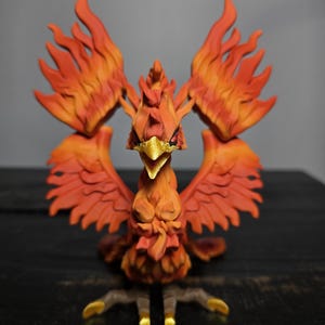 May include: A detailed, orange and red phoenix figurine with outstretched wings. The bird has a golden beak and talons, and its feathers are sculpted to resemble flames. The figurine is set against a dark background.