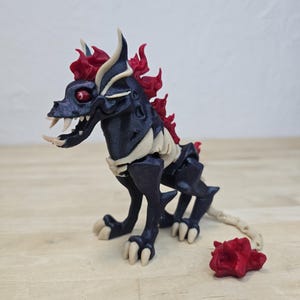 May include: A 3D-printed fantasy creature figurine. The figure is primarily black with bone-colored accents and red flame-like details. It has sharp teeth, red eyes, and a tail. A detached red piece sits nearby.