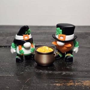May include: Two St. Patrick's Day gnome figurines with a pot of gold. One gnome wears a black hat and scarf with a green shamrock. The other gnome wears a black top hat with a green shamrock. The pot is bronze and filled with gold.