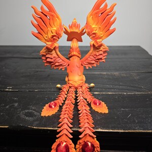 May include: A vibrant orange and yellow phoenix sculpture with outstretched wings and a crown. The bird's tail feathers feature red jewel-like accents. The sculpture is set against a dark wooden surface.