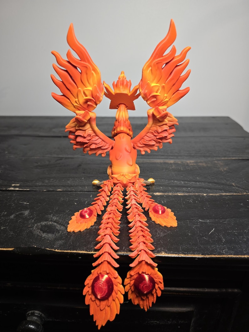 May include: A vibrant orange and yellow phoenix sculpture with outstretched wings and a crown. The bird's tail feathers feature red jewel-like accents. The sculpture is set against a dark wooden surface.