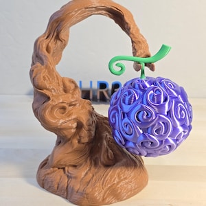 May include: A brown, tree-like sculpture supporting a purple, spherical fruit with a green stem. The fruit has a detailed, swirling pattern. The sculpture is set on a light-colored wooden surface.