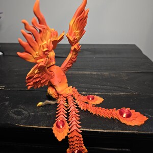 May include: A detailed orange and yellow phoenix sculpture with outstretched wings and tail feathers. The sculpture has a fiery appearance, with red accents on the tail feathers. The phoenix is perched on a dark wooden surface.