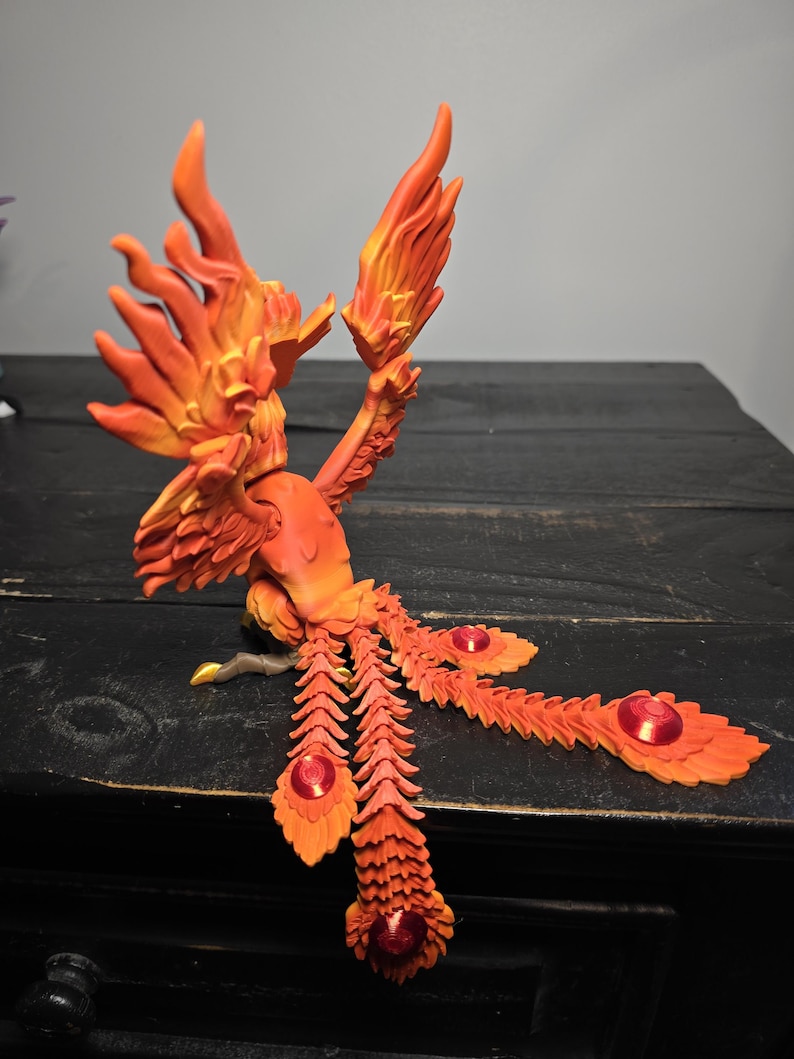 May include: A detailed orange and yellow phoenix sculpture with outstretched wings and tail feathers. The sculpture has a fiery appearance, with red accents on the tail feathers. The phoenix is perched on a dark wooden surface.