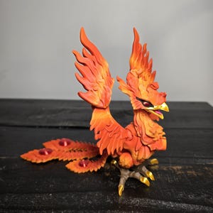 May include: A detailed figurine of a phoenix, featuring vibrant orange and red hues. The mythical bird has outstretched wings and a flowing tail with red accents. The phoenix stands on a dark surface, showcasing its golden talons and beak.