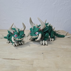 May include: Two green dragon figurines with cream-colored horns, teeth, and claws. The dragons have red eyes and spiky details along their backs. One dragon is smaller than the other. The figurines are on a wooden surface.