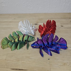 May include: Four articulated fish sculptures in white, red, green/gold, and purple/blue hues. The fish have detailed, fan-like fins and a segmented body design. They are arranged on a light wood surface.