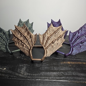 May include: A collection of decorative, wing-shaped headbands in various colors, including bronze, purple, and green. Each wing has a textured, scale-like pattern and is attached to a brown headband base. The headbands are arranged on a dark wooden surface.